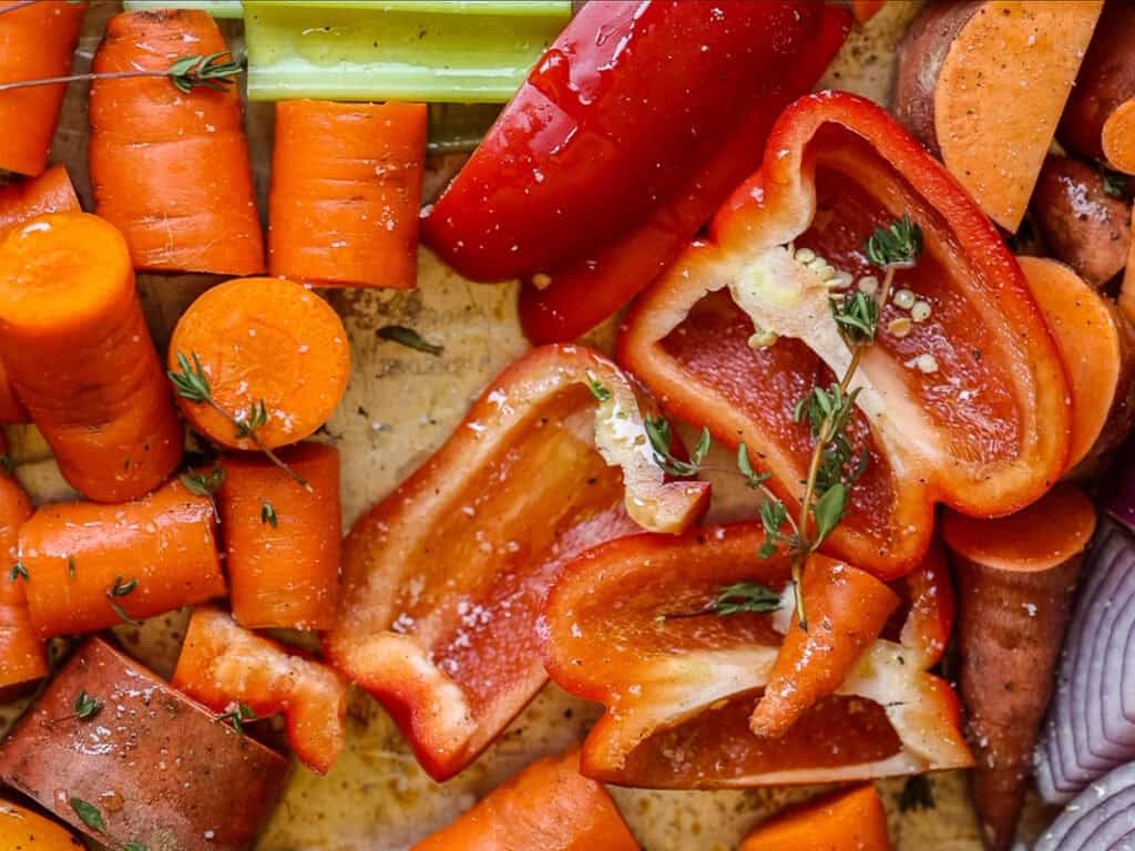 Chopped vegetables including sliced carrots, red bell peppers, and sweet potatoes are seasoned with herbs on a tray, ready for roasting.